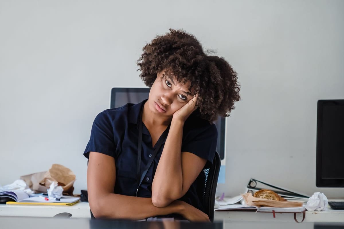 Femme assise à son bureau, la tête appuyée sur sa main, l'air las, au milieu de papiers en désordre