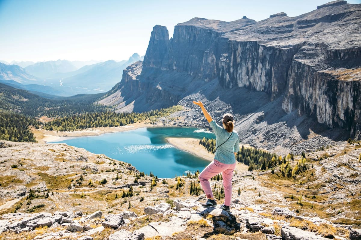 Une femme au sommet d'une falaise, bras levé, face à un lac de montagne